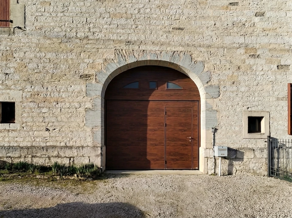 Façade en pierre calcaire rustique avec une porte de garage sectionnelle à relevage vertical, finition bois noyer foncé, intégrée dans une arche en pierre de taille
