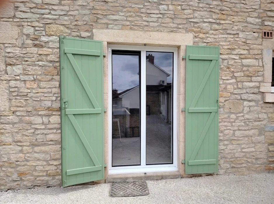 Volet coulissant en aluminium gris clair installé en façade extérieure d'une maison, partiellement ouvert devant une baie vitrée à deux vantaux avec rideau blanc