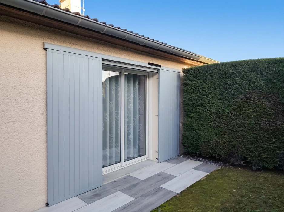 Volet coulissant en aluminium gris clair installé en façade extérieure d'une maison, partiellement ouvert devant une baie vitrée à deux vantaux avec rideau blanc