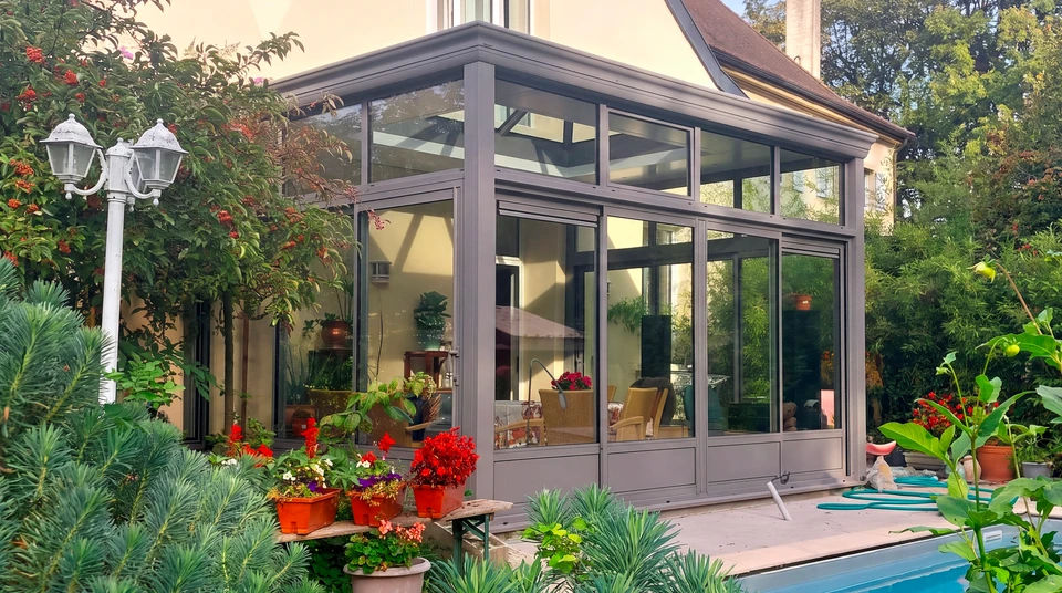 Véranda moderne vue de l'intérieur, salon de jardin gris anthracite, toiture vitrée à deux pans avec faîtage central, ossature aluminium laqué gris anthracite, vitrage feuilleté anti-UV, menuiseries coulissantes grande hauteur, parois latérales entièrement vitrées, raccordement à l'habitation principale par baie coulissante, sol carrelé clair